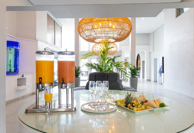 Bright hotel lobby refreshment table with two juice dispensers, champagne flutes, fruit platter and sandwiches beneath woven pendant lights and plants