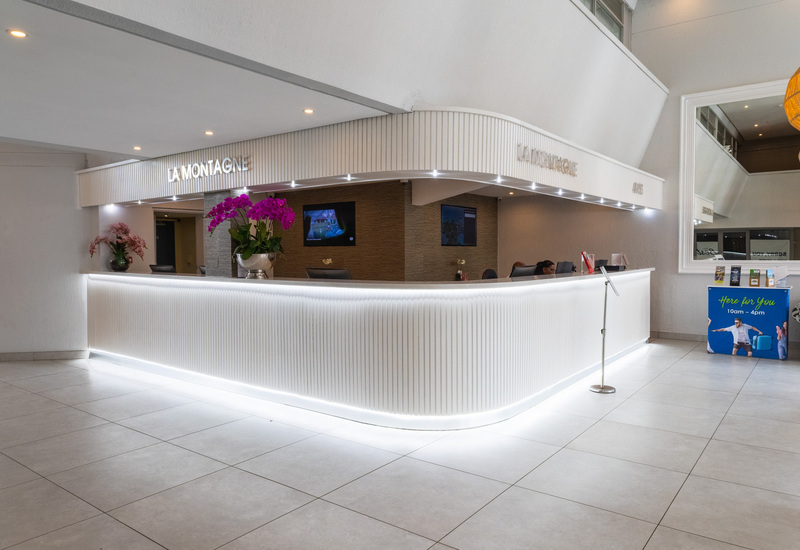Bright, modern hotel reception with curved backlit white desk labeled 'LA MONTAGNE', purple orchids, staff at check-in and brochure stand.