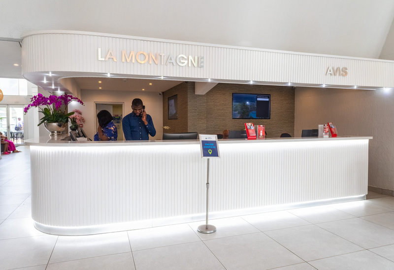 Modern La Montagne reception with curved white desk, staff on phone, orchids, 'LA MONTAGNE' signage and online check‑in sign.