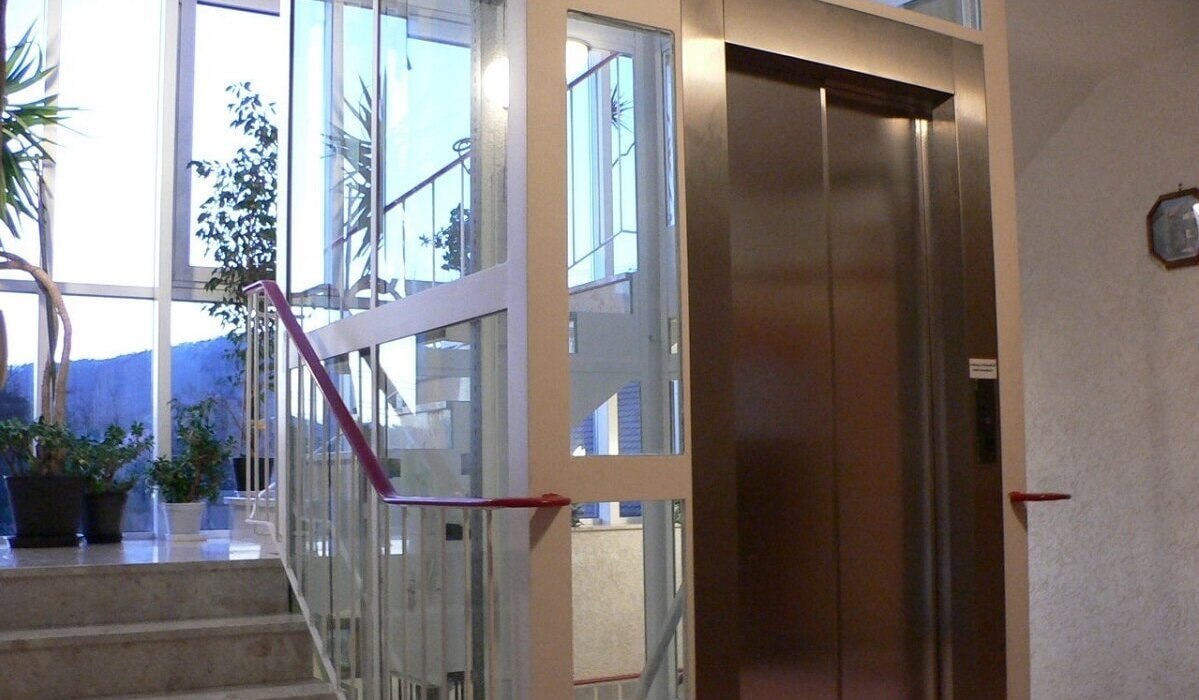 Moderner Hotellift im hellen Treppenhaus mit Glasfront und Pflanzen.