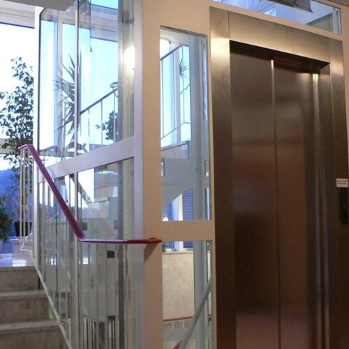 Moderner Hotellift im hellen Treppenhaus mit Glasfront und Pflanzen.