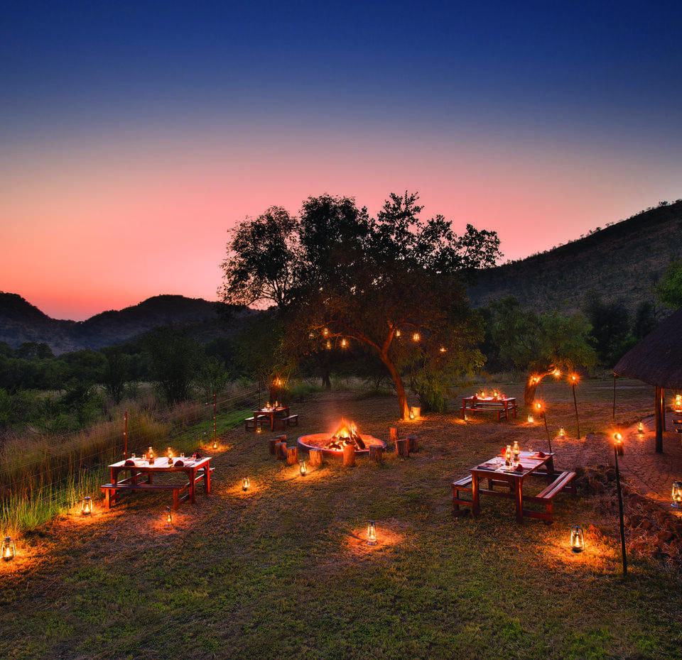 Intimate lantern-lit safari dining with wooden tables around a central campfire near a thatched lodge, set against a pink dusk sky and rolling hills.