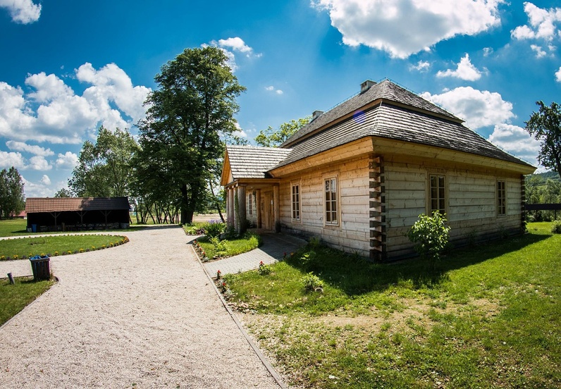 Tradycyjny drewniany domek z dachem krytym gontem, otoczony zielonym trawnikiem i drzewami pod błękitnym niebem.