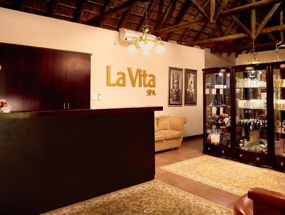 Warm spa reception with 'La Vita SPA' sign, wood desk, product display cabinets, plush seating and thatched ceiling.