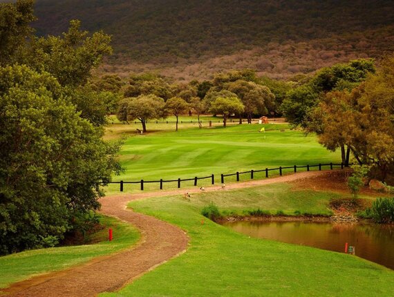 Lush Magalies Park golf course with manicured fairway and flag, winding cart path, water hazard and tree-lined mountain backdrop.