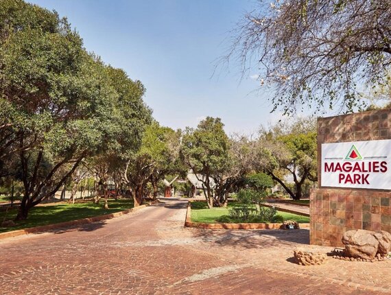 Tree-lined brick driveway leading to Magalies Park entrance with tiled stone sign, shaded lawns and rustic lodge buildings beyond.