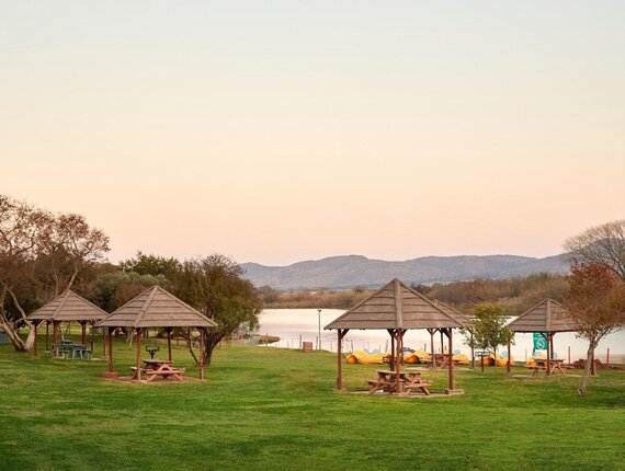 Lakeside resort lawn with wooden picnic gazebos, benches and yellow kayaks on the shore, calm water and low hills at sunset.
