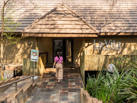 Guest entering La Vita Spa & Hair Studio on shaded tiled path, thatched-roof entrance framed by bamboo and greenery.