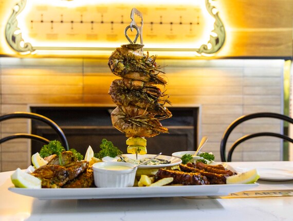 Vertical skewer of grilled prawns over a platter with sauces, fries and lemon wedges in a warm hotel bistro