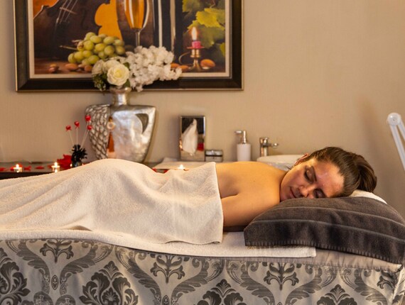 Guest reclining on massage table under a white towel in a softly lit spa room with candles, floral vase and artwork.