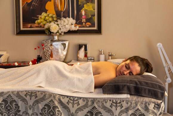 Guest reclining on massage table under a white towel in a softly lit spa room with candles, floral vase and artwork.