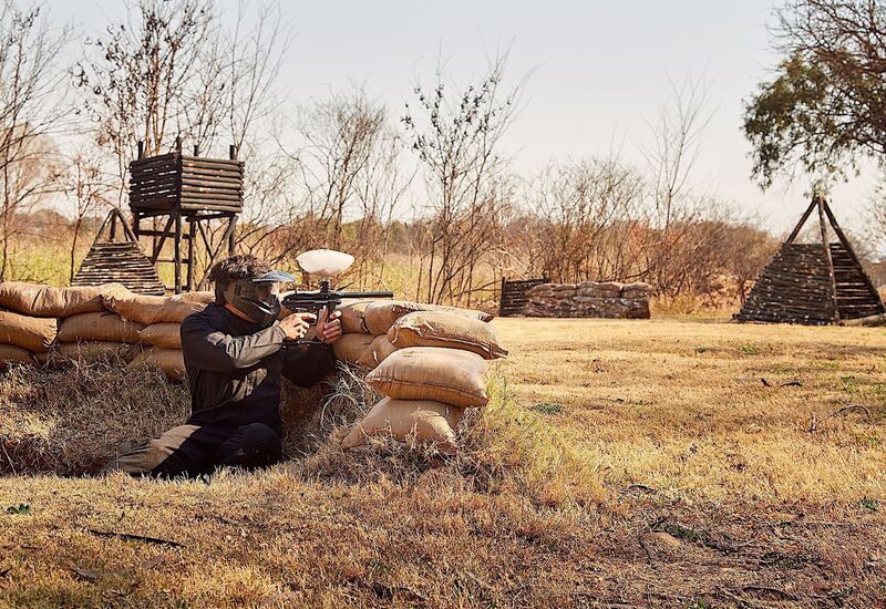 Guest in protective mask firing a paintball marker from sandbag bunker on an outdoor paintball field with wooden towers