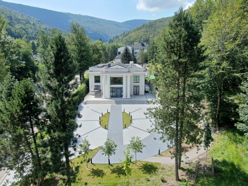 Biała willa hotelowa z kolumnami, tarasem z parasolem i okrągłym podjazdem, otoczona sosnowym lasem i górami
