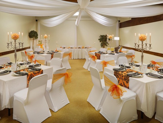 Elegant wedding reception room with draped white ceiling, round and rectangular tables, white chair covers tied with orange bows and silver candelabra