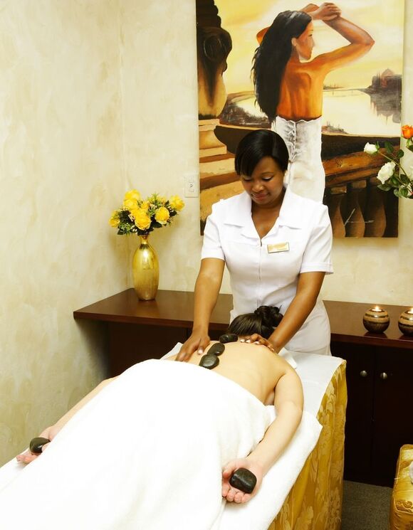 Therapist performing hot-stone back massage on draped guest in serene spa room with candles, fresh roses and soft lighting.