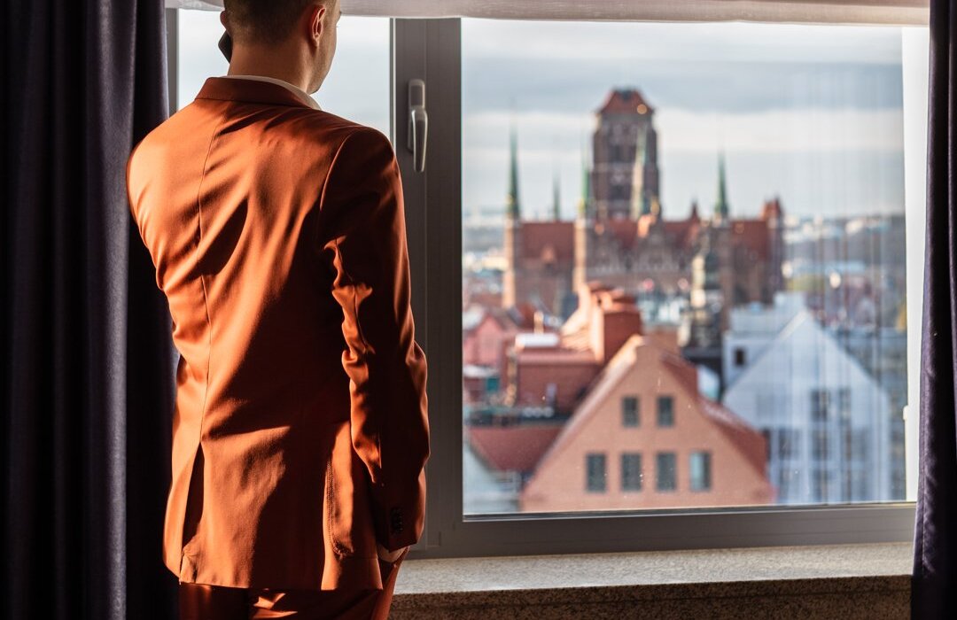 Mężczyzna w czerwonym garniturze podziwiający przez okno widok na gdańskie Stare Miasto.