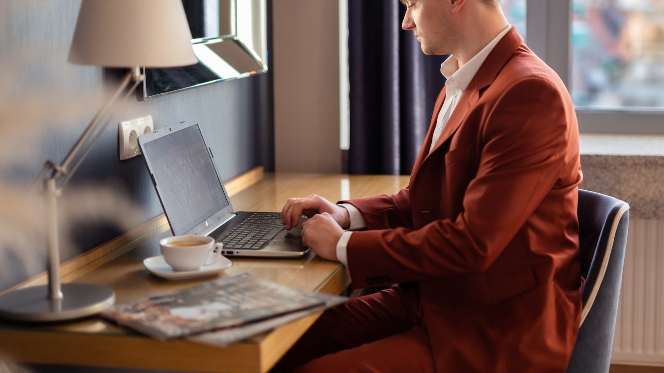 Mężczyzna w brązowym garniturze pracujący na laptopie przy hotelowym biurku.