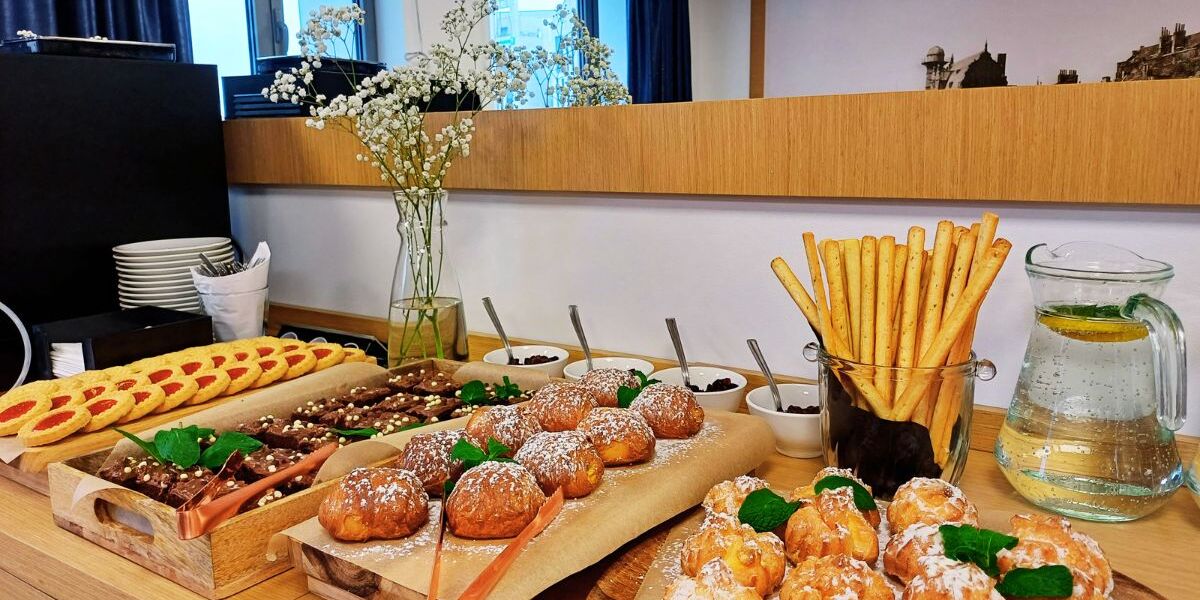Bufet z różnorodnymi wypiekami, grissini i dzbankiem wody z cytryną w jasnej hotelowej sali śniadaniowej.