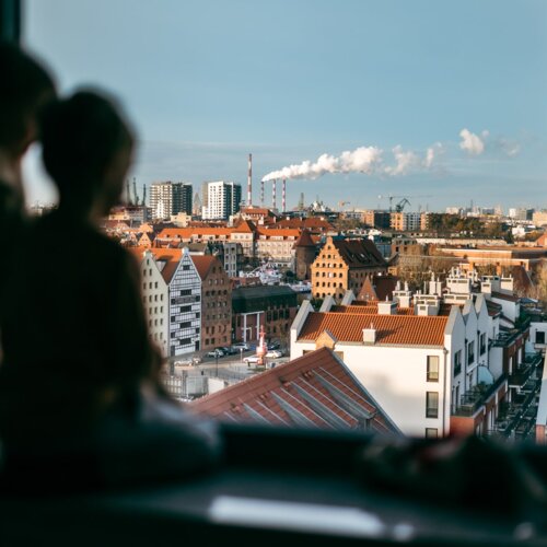 Postać siedząca na parapecie okna i wpatrująca się w widok na Stare Miasto w Gdańsku.