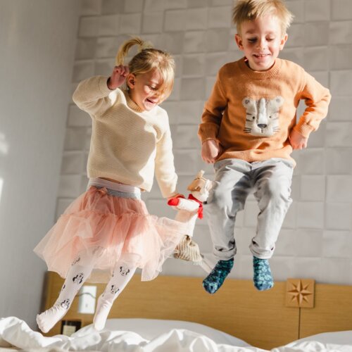 Dziewczynka w różowej spódniczce i chłopiec w pomarańczowej bluzie radośnie skaczą po łóżku.