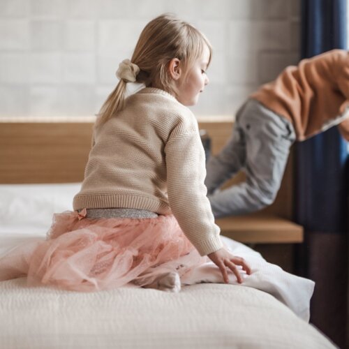 Uśmiechnięta dziewczynka w beżowym sweterku i różowej spódniczce siedząca na łóżku.