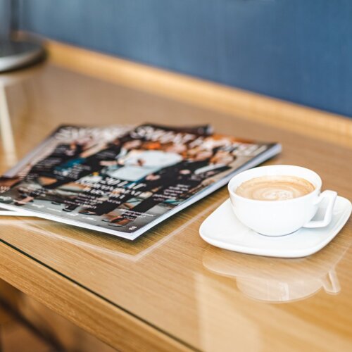 Biała filiżanka kawy ze spodkiem i kolorowe czasopisma leżące na biurku.