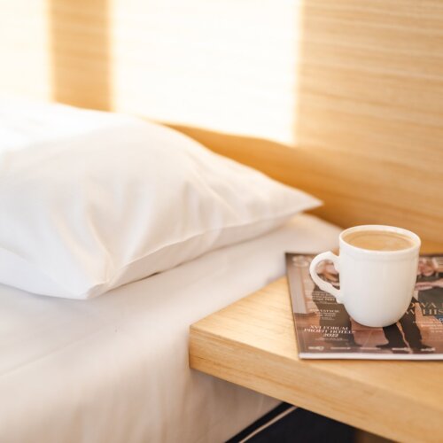 Biały kubek z kawą, leżący na kolorowym czasopiśmie na stoliku nocnym przy łóżku.