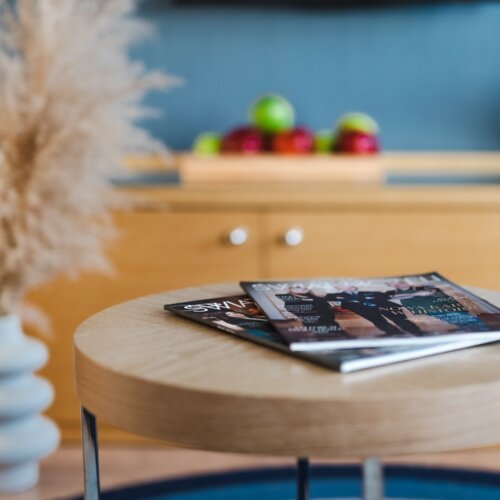 Kolorowe czasopisma leżące na okrągłym stoliku kawowym z trawą pampasową i jabłkami w tle.