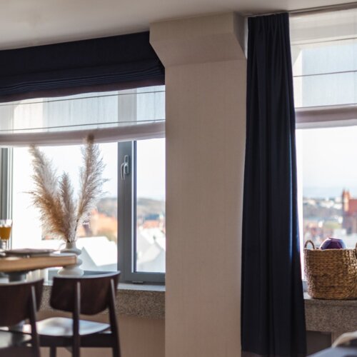 Pokój ze stolikiem i dwoma krzesłami barowymi i dwoma dużymi oknami z widokiem na miasto.