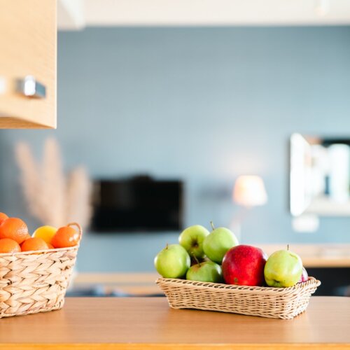 Czerwone i zielone jabłka w wiklinowym koszyku stojące na blacie aneksu kuchennego.