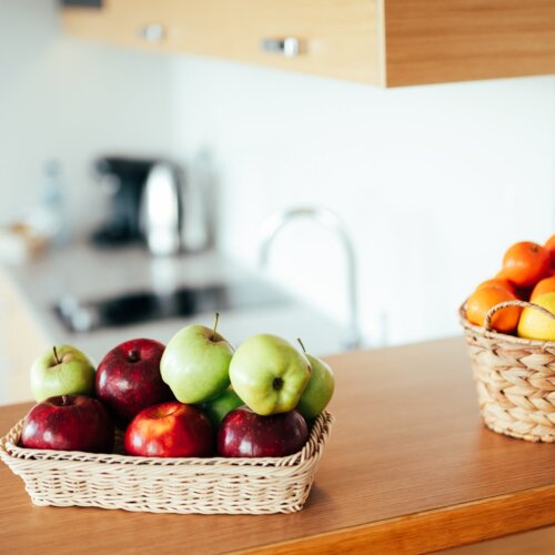 Czerwone i zielone jabłka w wiklinowym koszyku stojące na blacie aneksu kuchennego.