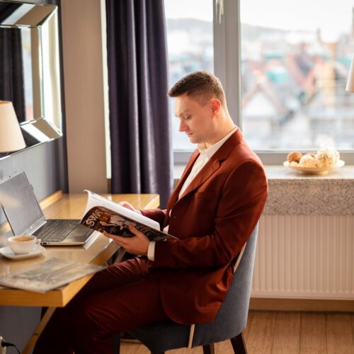 Mężczyzna w brązowym garniturze czyta czasopismo przy biurku w pokoju hotelowym.