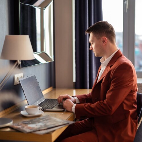 Mężczyzna w brązowym garniturze pracujący na laptopie przy hotelowym biurku.