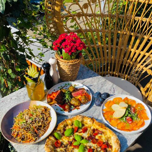 Taras restauracyjny z kamiennym stołem, na którym podano pizzę z bazylią, stek, sałatkę i zimny napój z owocami.