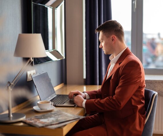 Mężczyzna w brązowym garniturze pracujący na laptopie przy hotelowym biurku.