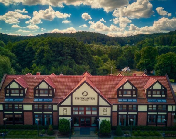 Dwupiętrowy budynek z czerwoną dachówką. W tle widnieje wspaniały las oraz pogodne niebo .