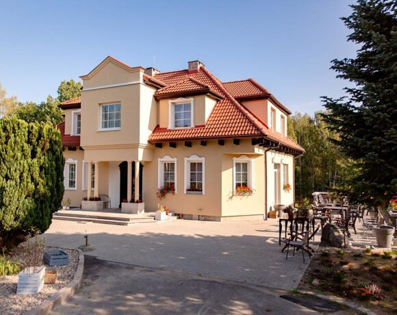 Mały dwupiętrowy dworek z jasnymi ścianami i czerwoną dachówką otoczony drzewami i krzewami.