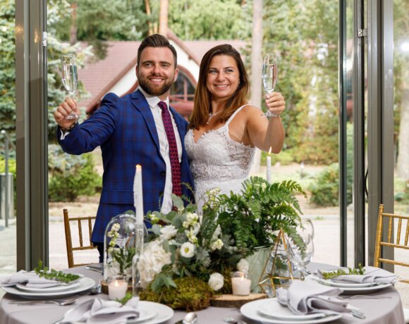 Mężczyzna w granatowej marynarce oraz kobieta w białej sukni wznoszą toast przy nakrytym stole.