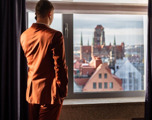Mężczyzna w czerwonym garniturze podziwiający przez okno widok na gdańskie Stare Miasto.