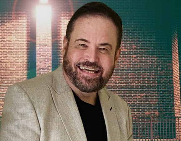 Smiling man in a beige blazer and black shirt by a lit brick exterior wall and metal railing, warm evening ambiance