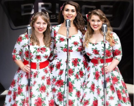 Vintage vocal trio in matching 1950s floral dresses and red belts singing at a live hotel stage with retro microphones
