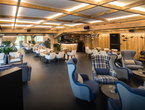 Cozy hotel lounge with wooden-beam ceiling, bar, white dining chairs, blue plaid armchairs, plants and warm lighting.