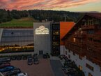 Hotel Meta Resort & Spa mit leuchtendem Logo, Glasfassade mit Blick auf Hallenbad, angrenzender Holzchalet-Flügel und bewaldete Hänge bei Sonnenuntergang
