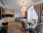 Modern hotel suite living area with wooden kitchenette, sofa, round dining table for four, large curtained window and pendant light.
