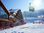 Chalet-style ski hotel with glass façade, wooden balconies and yellow chairlift carrying skiers above a sunlit snowy slope.