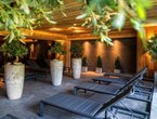 Indoor-Spa-Lounge mit Holzbalkendecke, Reihe dunkler Liegen, großen Steinpflanzgefäßen mit Olivenbäumen und warmem Licht