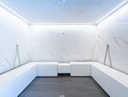 Spa-Raum mit marmorverkleideten Wänden, U-förmigen weißen Bänken, zwei Handbrausen und eingelassener LED-Deckenbeleuchtung.