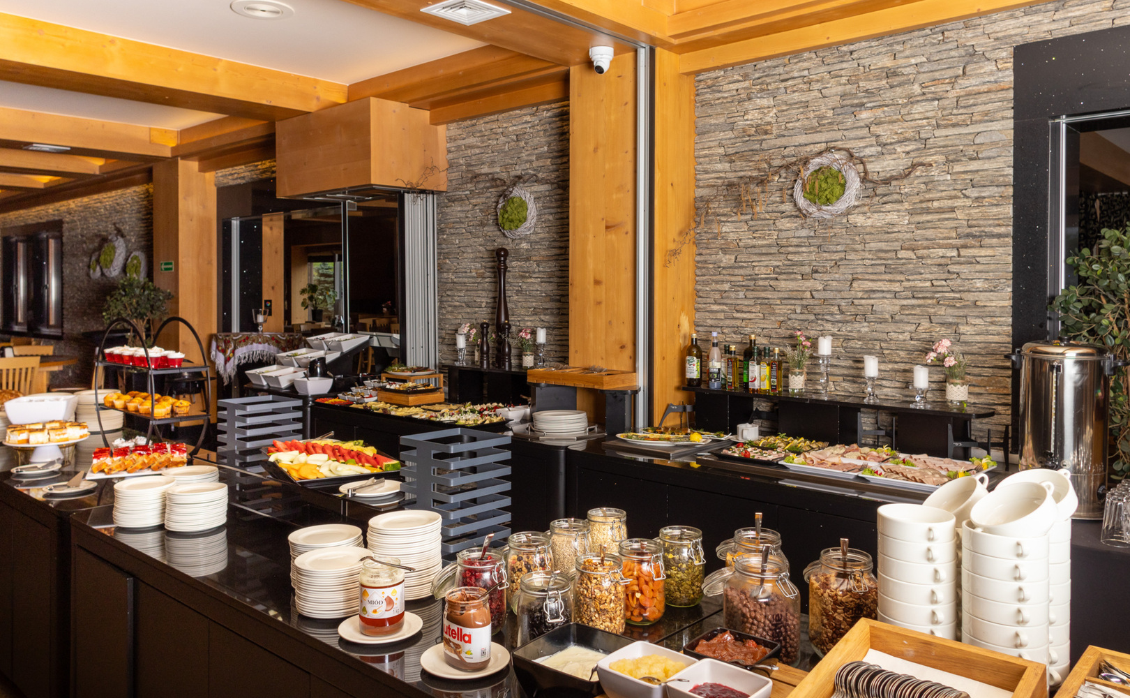Hotel-Frühstücksbuffet mit frischen Obstplatten, Gebäck, Gläsern mit Müsli und Aufstrichen, gestapelten Tellern, Holzbalken und Natursteinwand