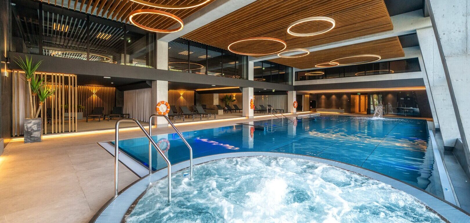 Überdachtes Hotel-Spa mit sprudelndem Whirlpool im Vordergrund, langem Schwimmbecken, Liegen, Holzlamellendecke und runden Hängeleuchten.