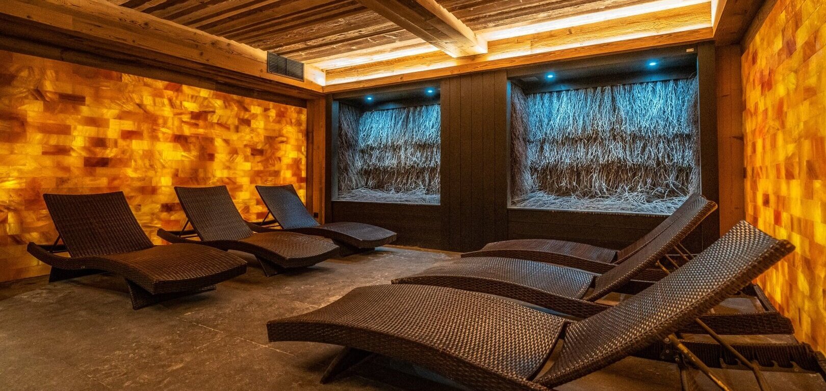 Entspannungsraum im Spa mit beleuchteter Himalaya-Salzwand, geflochtenen Liegen, Schilfnischen mit blauer Beleuchtung und warmen Holzdeckenbalken.